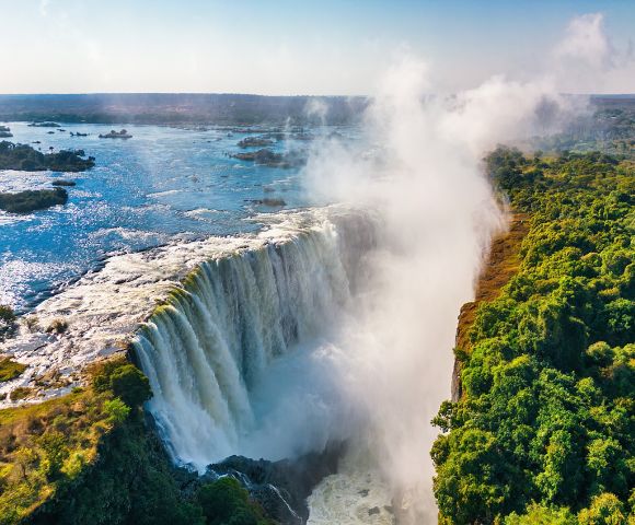 A majestic waterfall cascades into a gorge, with mist rising into a clear blue sky. Lush green foliage borders the falls on the right, adding contrast.
