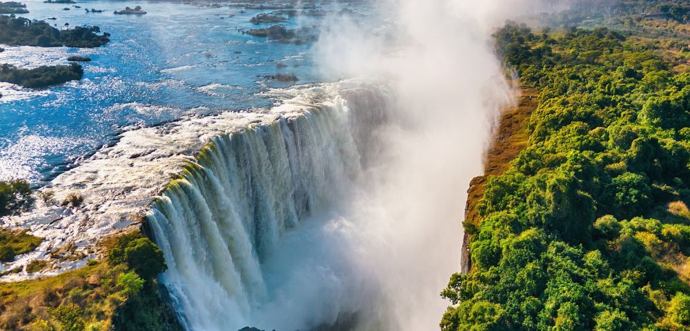 A majestic waterfall cascades into a gorge, with mist rising into a clear blue sky. Lush green foliage borders the falls on the right, adding contrast.