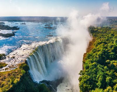 A majestic waterfall cascades into a gorge, with mist rising into a clear blue sky. Lush green foliage borders the falls on the right, adding contrast.