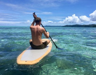 A person paddles on a stand-up paddleboard in clear, turquoise ocean water under a bright blue sky. Green hills are visible in the distance.