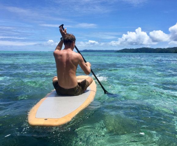 A person paddles on a stand-up paddleboard in clear, turquoise ocean water under a bright blue sky. Green hills are visible in the distance.