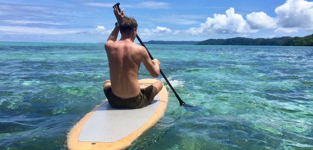 A person paddles on a stand-up paddleboard in clear, turquoise ocean water under a bright blue sky. Green hills are visible in the distance.