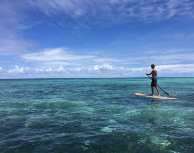A person stands on a paddleboard over clear, turquoise ocean water under a bright blue sky with fluffy clouds; the scene feels tranquil and carefree.