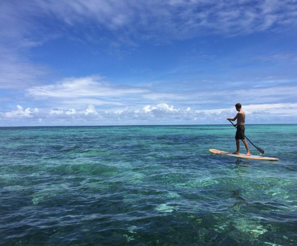A person stands on a paddleboard over clear, turquoise ocean water under a bright blue sky with fluffy clouds; the scene feels tranquil and carefree.