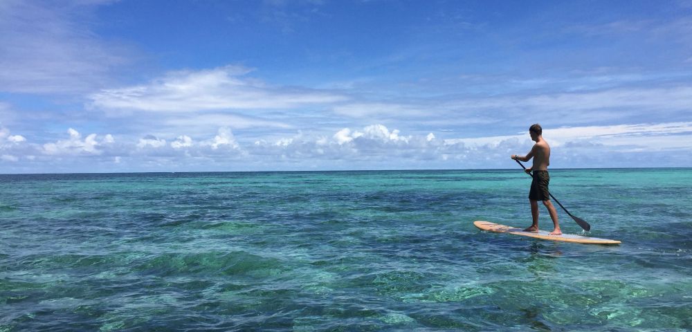A person stands on a paddleboard over clear, turquoise ocean water under a bright blue sky with fluffy clouds; the scene feels tranquil and carefree.