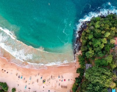 Aerial view of a tropical beach with turquoise waves meeting sandy shore, scattered colorful boats, lush green forest bordering, evoking tranquility and adventure.