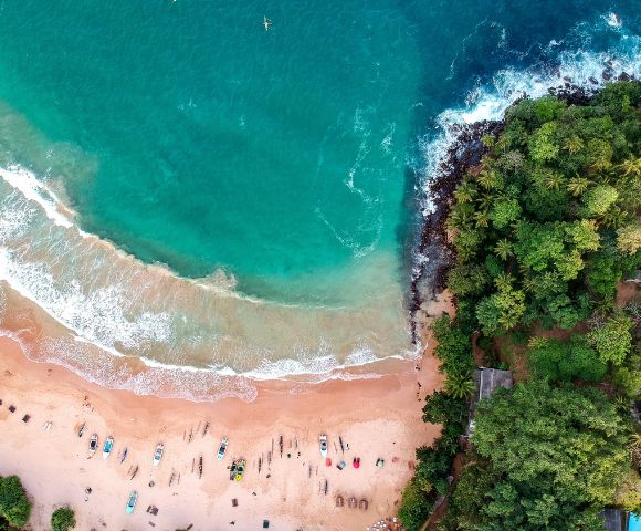 Aerial view of a tropical beach with turquoise waves meeting sandy shore, scattered colorful boats, lush green forest bordering, evoking tranquility and adventure.