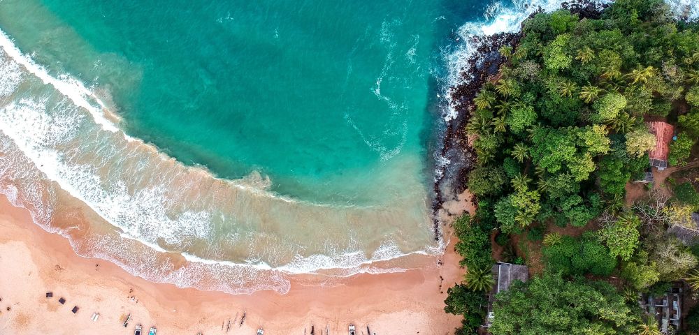 Aerial view of a tropical beach with turquoise waves meeting sandy shore, scattered colorful boats, lush green forest bordering, evoking tranquility and adventure.