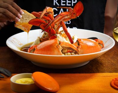 A plate of vibrant steamed crab claws with sauce, surrounded by bread, on an orange tablecloth. A hand dips bread in a sauce, conveying a warm dining atmosphere.