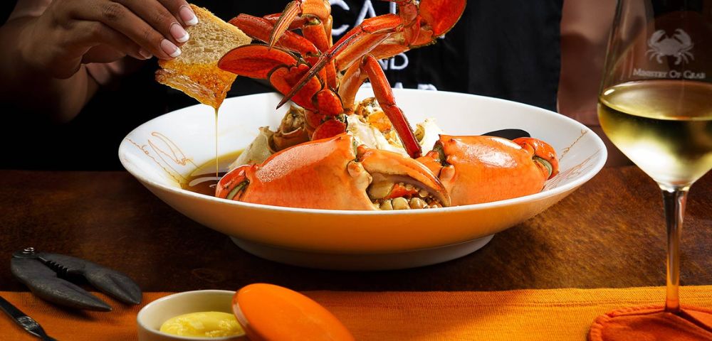 A plate of vibrant steamed crab claws with sauce, surrounded by bread, on an orange tablecloth. A hand dips bread in a sauce, conveying a warm dining atmosphere.