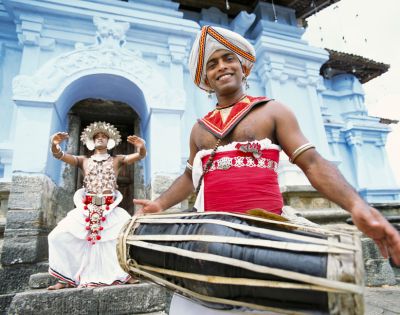 A man in traditional Sri Lankan attire plays a drum with a smile, while another in ornate costume dances behind him in front of a blue temple entrance.