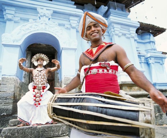A man in traditional Sri Lankan attire plays a drum with a smile, while another in ornate costume dances behind him in front of a blue temple entrance.