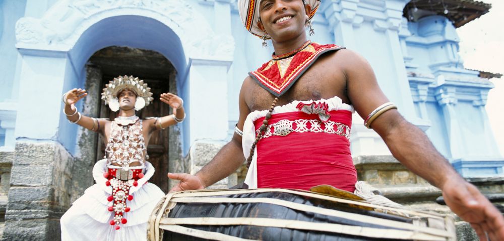 A man in traditional Sri Lankan attire plays a drum with a smile, while another in ornate costume dances behind him in front of a blue temple entrance.