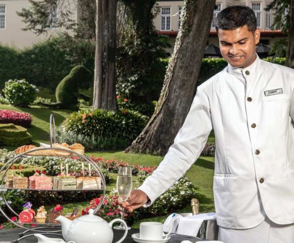 A waiter in a white uniform serves tea in a scenic garden. A tiered stand holds assorted pastries alongside a teapot and teacups, creating an elegant and serene atmosphere.