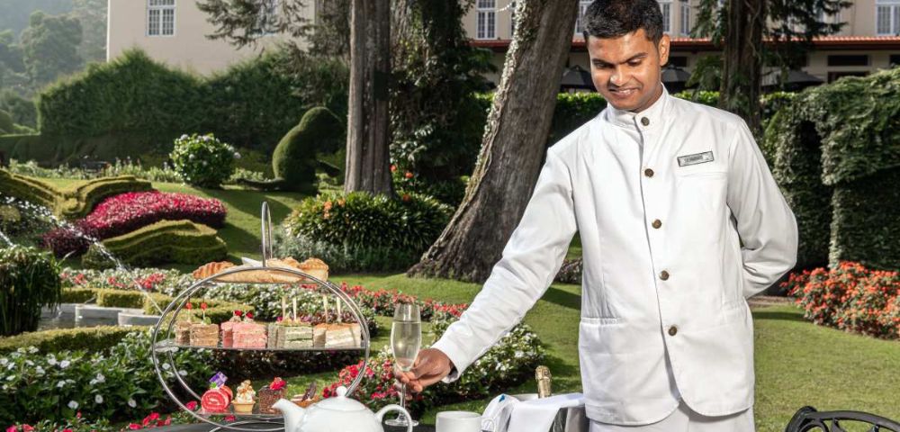A waiter in a white uniform serves tea in a scenic garden. A tiered stand holds assorted pastries alongside a teapot and teacups, creating an elegant and serene atmosphere.