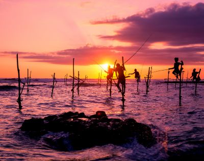 Silhouetted fishermen balance on stilts in the ocean at sunset, with vivid orange and purple hues in the sky, creating a serene and timeless scene.