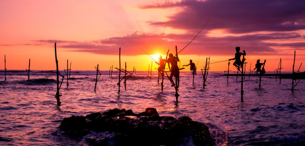Silhouetted fishermen balance on stilts in the ocean at sunset, with vivid orange and purple hues in the sky, creating a serene and timeless scene.