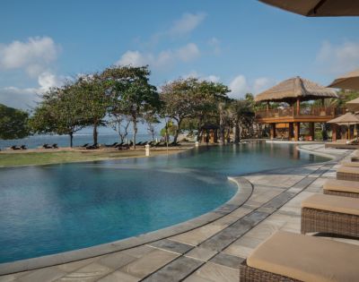 Luxurious poolside view with wicker loungers, surrounded by tropical trees and a thatched-roof pavilion, exuding relaxation and a serene ocean backdrop.
