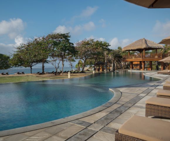 Luxurious poolside view with wicker loungers, surrounded by tropical trees and a thatched-roof pavilion, exuding relaxation and a serene ocean backdrop.