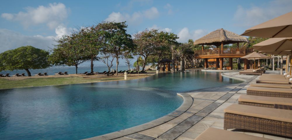 Luxurious poolside view with wicker loungers, surrounded by tropical trees and a thatched-roof pavilion, exuding relaxation and a serene ocean backdrop.