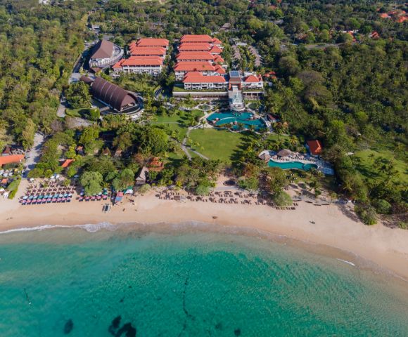 Aerial view of a beachfront resort with red-roofed buildings, lush greenery, and a large pool. Sandy beach and turquoise ocean in the foreground. Serene atmosphere.