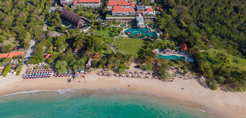 Aerial view of a beachfront resort with red-roofed buildings, lush greenery, and a large pool. Sandy beach and turquoise ocean in the foreground. Serene atmosphere.