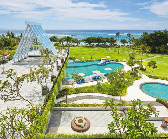 Luxurious beachfront resort with a glass chapel, lush gardens, and two turquoise pools overlooking the ocean. Sunny, tropical, and serene atmosphere.