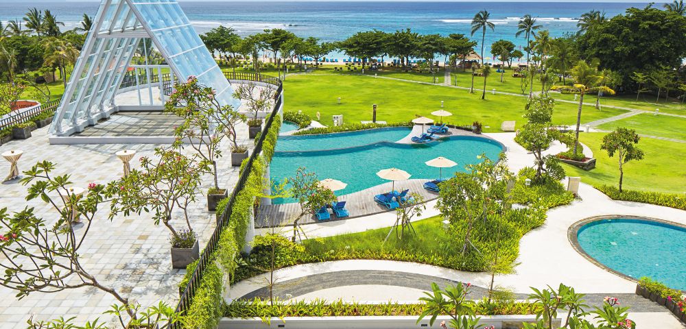 Luxurious beachfront resort with a glass chapel, lush gardens, and two turquoise pools overlooking the ocean. Sunny, tropical, and serene atmosphere.