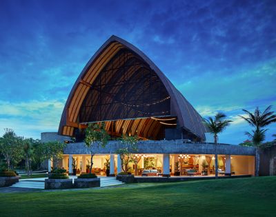 A stunning evening view of a modern, open-concept building with an arched, wooden roof, vibrant interior lighting, set against a twilight sky and lush greenery.