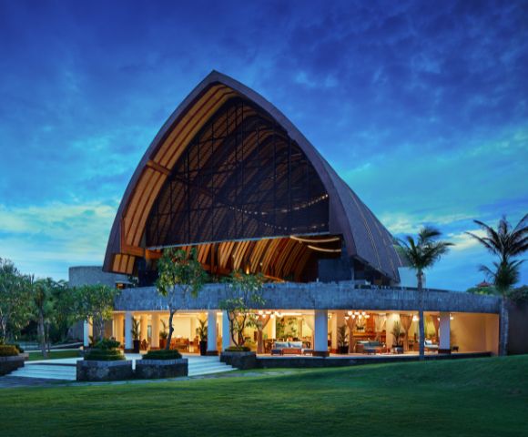 A stunning evening view of a modern, open-concept building with an arched, wooden roof, vibrant interior lighting, set against a twilight sky and lush greenery.