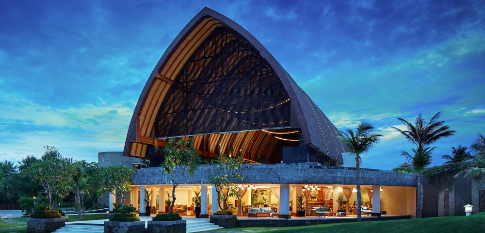 A stunning evening view of a modern, open-concept building with an arched, wooden roof, vibrant interior lighting, set against a twilight sky and lush greenery.