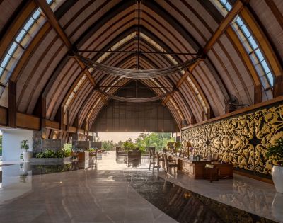 Majestic hotel lobby with a vaulted wooden ceiling, elegant decor, ornate gold wall panel, and lush greenery. Sunlight filters in, creating a serene, luxurious ambiance.