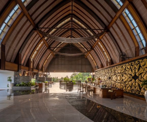 Majestic hotel lobby with a vaulted wooden ceiling, elegant decor, ornate gold wall panel, and lush greenery. Sunlight filters in, creating a serene, luxurious ambiance.
