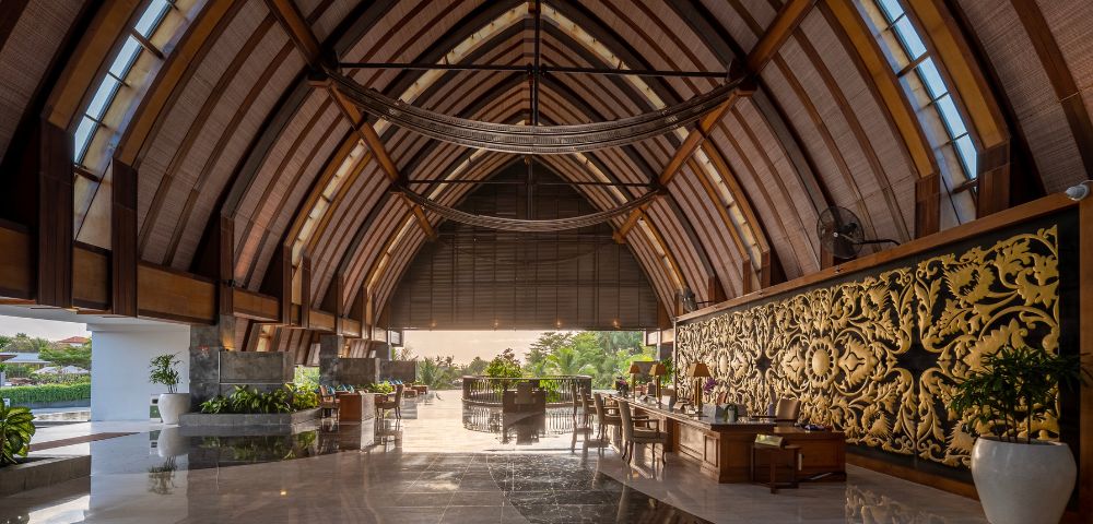Majestic hotel lobby with a vaulted wooden ceiling, elegant decor, ornate gold wall panel, and lush greenery. Sunlight filters in, creating a serene, luxurious ambiance.