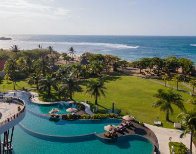 Aerial view of a tropical resort showing a large pool with umbrellas, sunbeds, and lush gardens. Palm trees lead to a sandy beach and calm ocean under clear skies.
