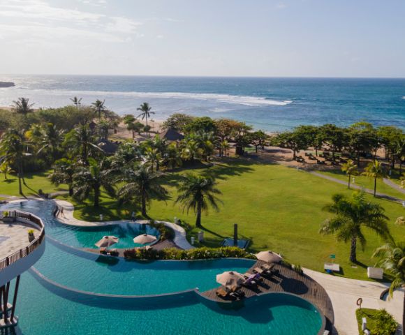 Aerial view of a tropical resort showing a large pool with umbrellas, sunbeds, and lush gardens. Palm trees lead to a sandy beach and calm ocean under clear skies.