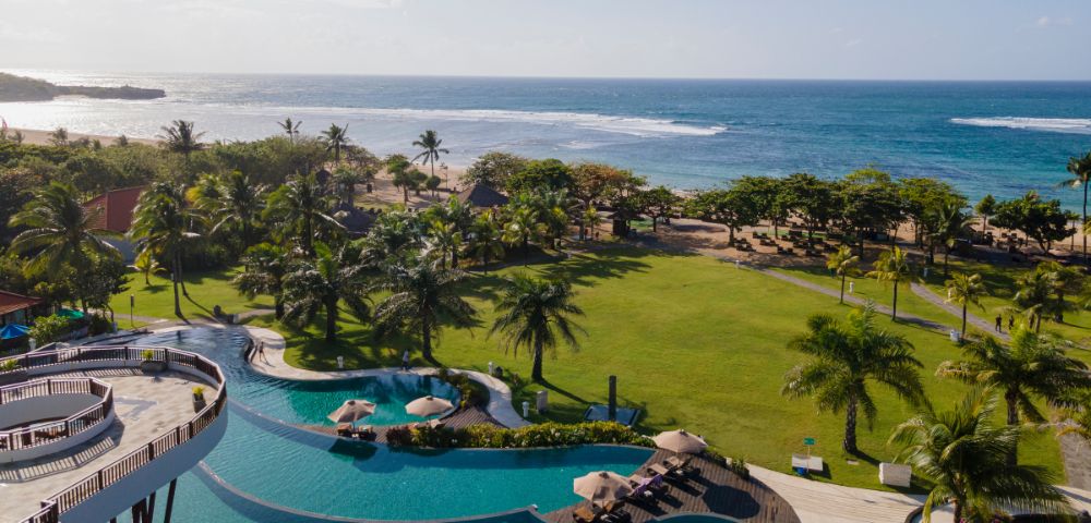 Aerial view of a tropical resort showing a large pool with umbrellas, sunbeds, and lush gardens. Palm trees lead to a sandy beach and calm ocean under clear skies.