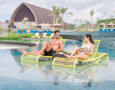 A man and woman relax on lime-green lounge chairs in a pool, enjoying drinks. The background features tropical plants and a large dome structure.