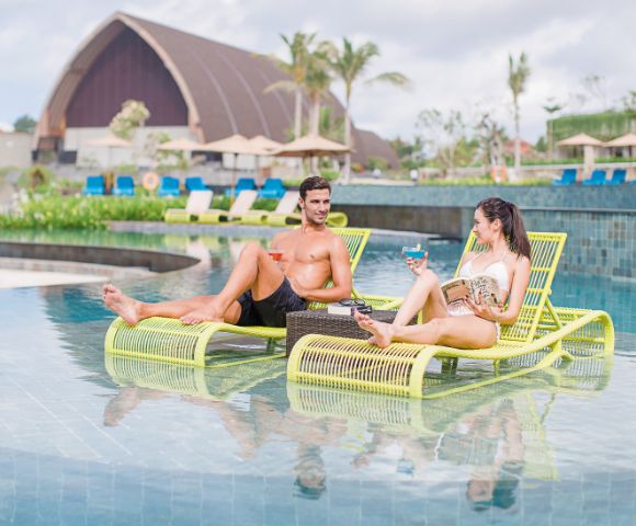 A man and woman relax on lime-green lounge chairs in a pool, enjoying drinks. The background features tropical plants and a large dome structure.