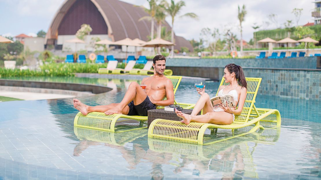 A man and woman relax on lime-green lounge chairs in a pool, enjoying drinks. The background features tropical plants and a large dome structure.