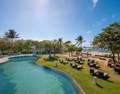 Tropical beachfront scene with a pool, lined with lounge chairs and palm trees. Clear blue sky and ocean create a relaxing and serene atmosphere.