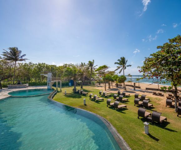 Tropical beachfront scene with a pool, lined with lounge chairs and palm trees. Clear blue sky and ocean create a relaxing and serene atmosphere.