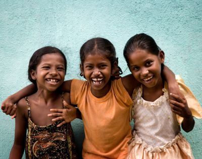 Three smiling children with arms around each other stand in front of a turquoise wall. They exude joy and friendship, wearing colorful, casual clothing.