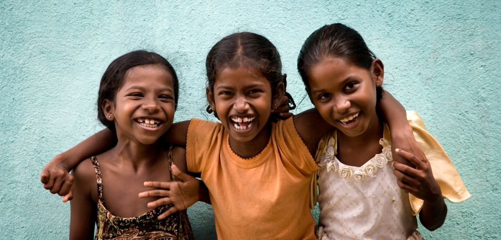 Three smiling children with arms around each other stand in front of a turquoise wall. They exude joy and friendship, wearing colorful, casual clothing.