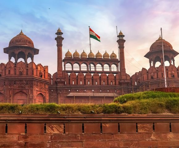 Historic Red Fort with red sandstone walls under the Indian flag, set against a vivid sunset sky. The scene conveys grandeur and national pride.
