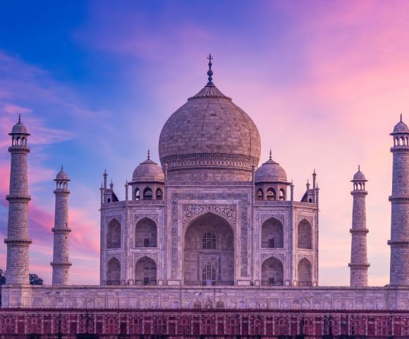The Taj Mahal against a vibrant sunset sky. The iconic white marble structure, with its central dome and four minarets, exudes a serene, majestic beauty.