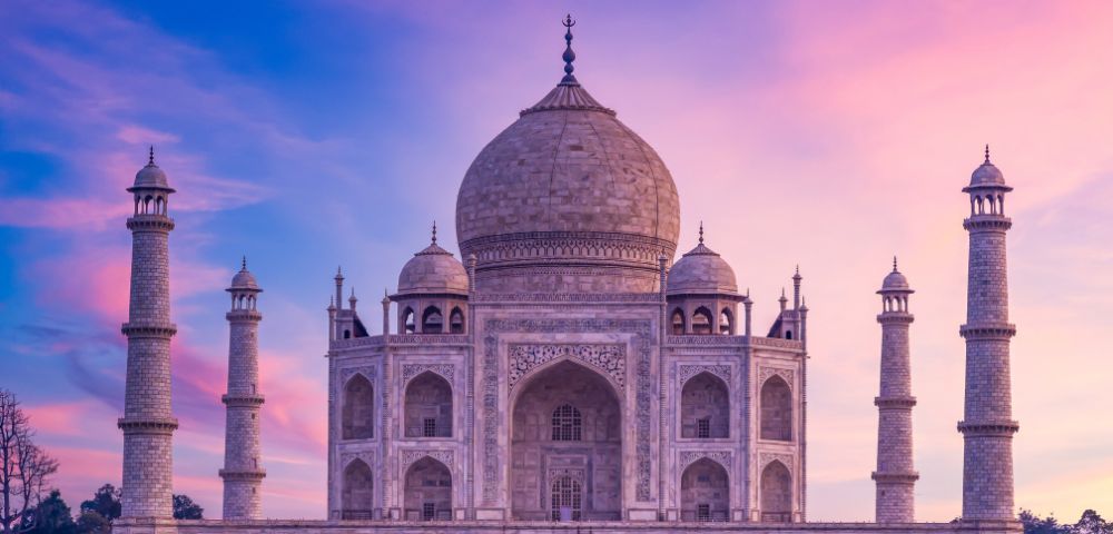 The Taj Mahal against a vibrant sunset sky. The iconic white marble structure, with its central dome and four minarets, exudes a serene, majestic beauty.