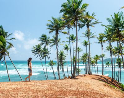A woman in shorts and a tank top walks on a cliff surrounded by tall palm trees, overlooking a vibrant blue ocean under a clear, sunny sky.