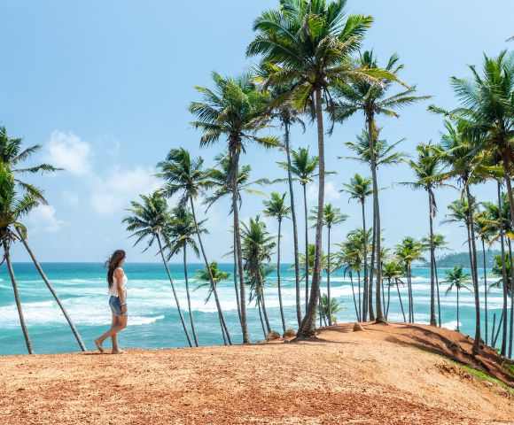 A woman in shorts and a tank top walks on a cliff surrounded by tall palm trees, overlooking a vibrant blue ocean under a clear, sunny sky.