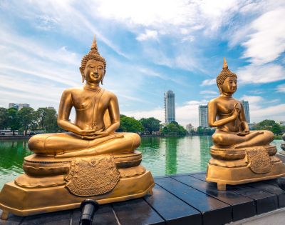 Two golden Buddha statues sit serenely by a tranquil lake, set against a backdrop of lush trees and modern skyscrapers under a partly cloudy sky.